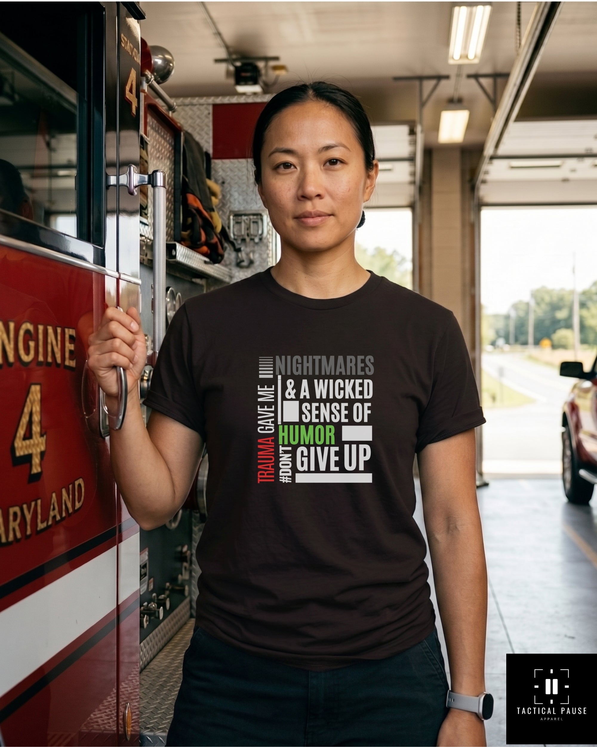 Female firefighter, in the fire station, wearing a dark gray t-shirt with text "Trauma gave me nightmare" in front of a black truck. Tactical Pause Apparel