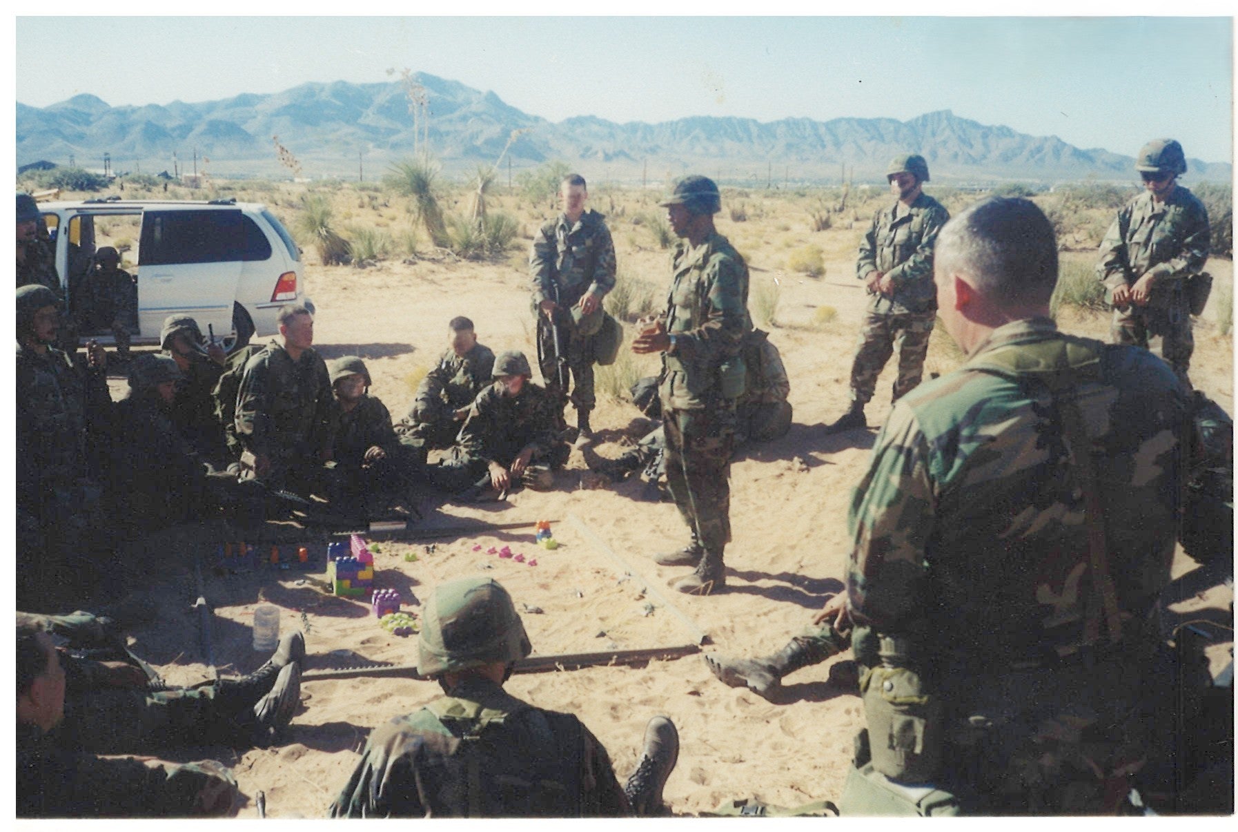 Group of soldiers in a desert setting with mountains in the background, giving a briefing