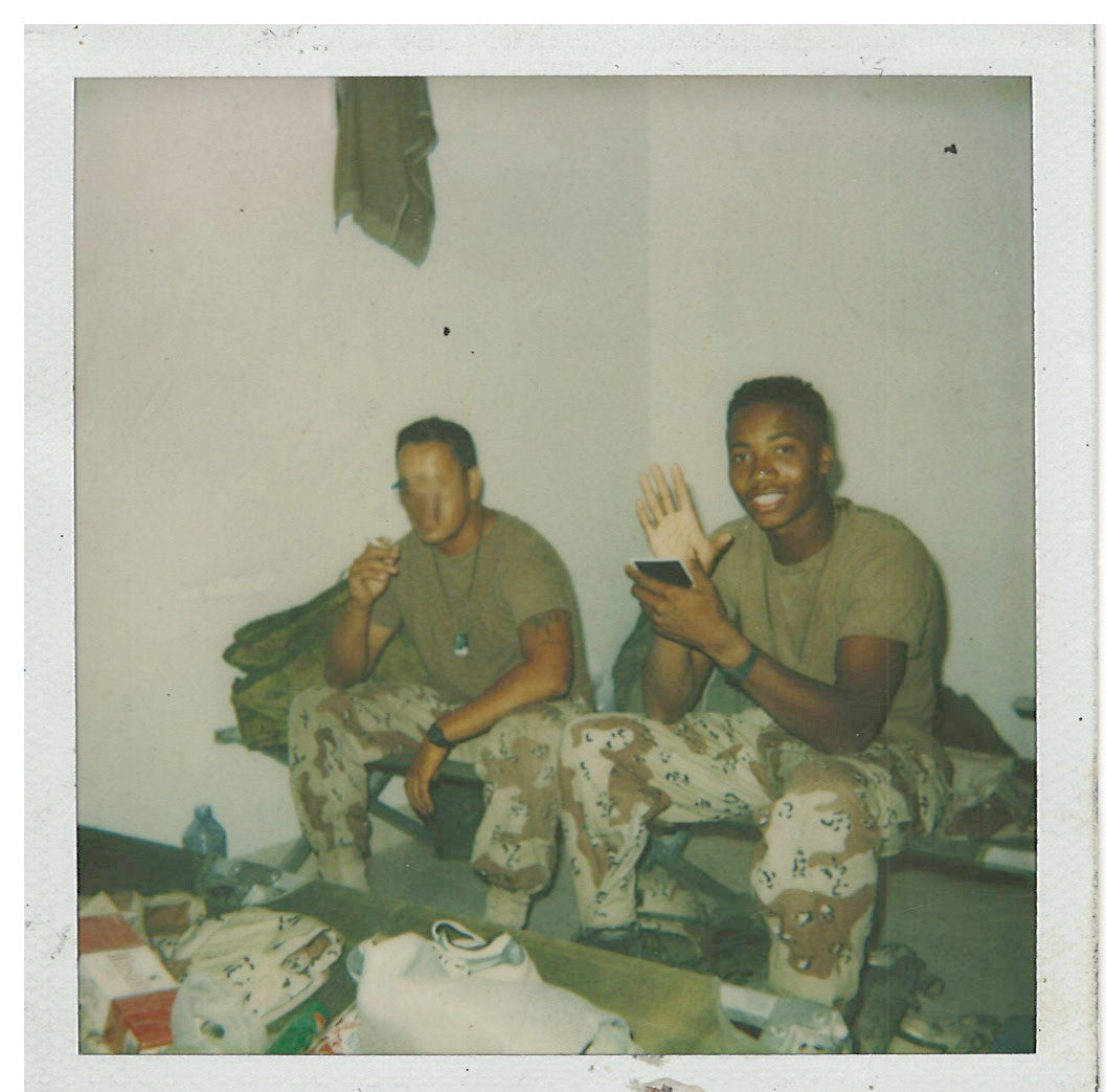 Two individuals in military uniforms sitting on a cot in a room, during a deployment with various items around.