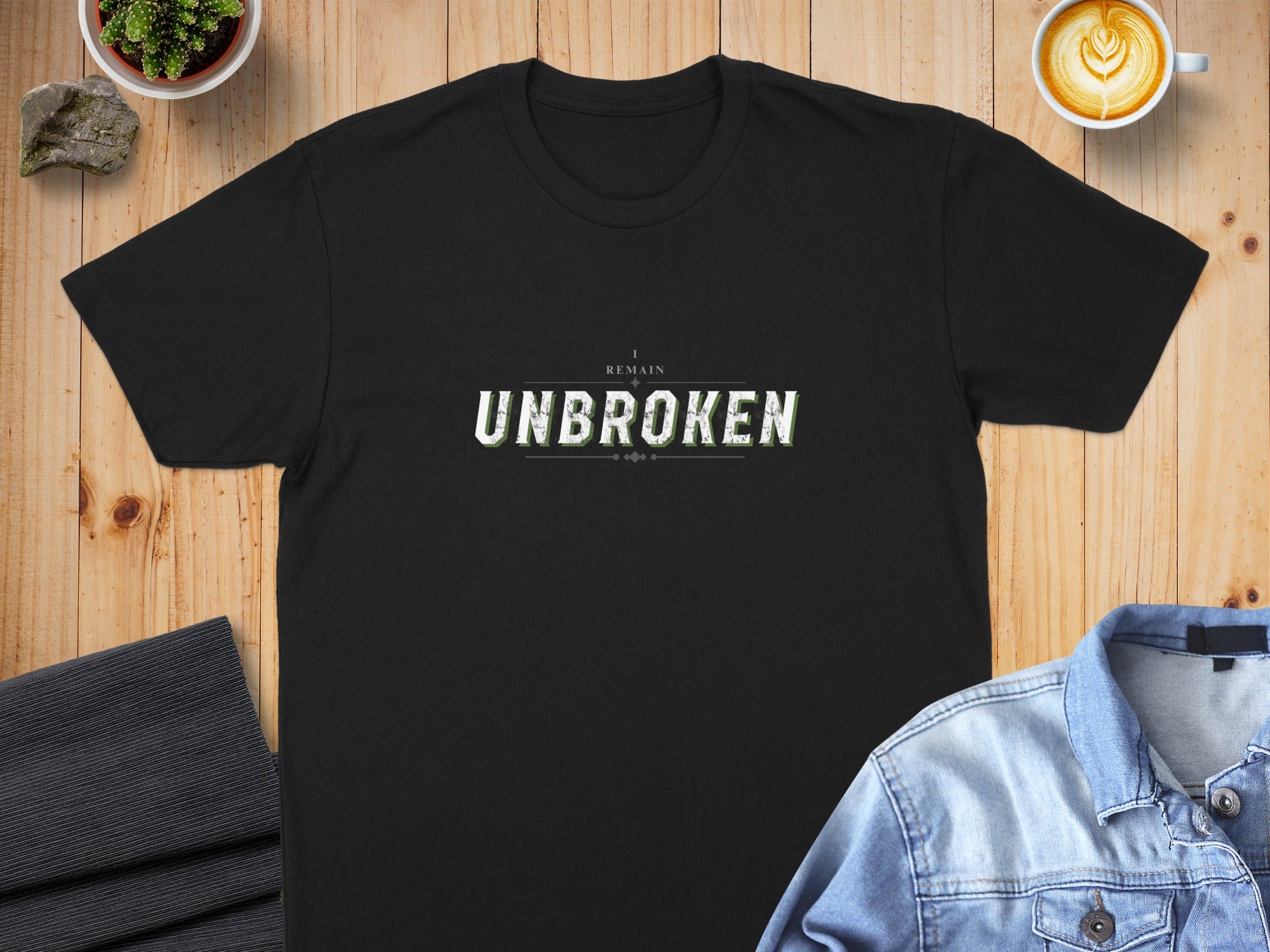 Black t-shirt with "I REMAIN UNBROKEN" text, displayed on a wooden surface.