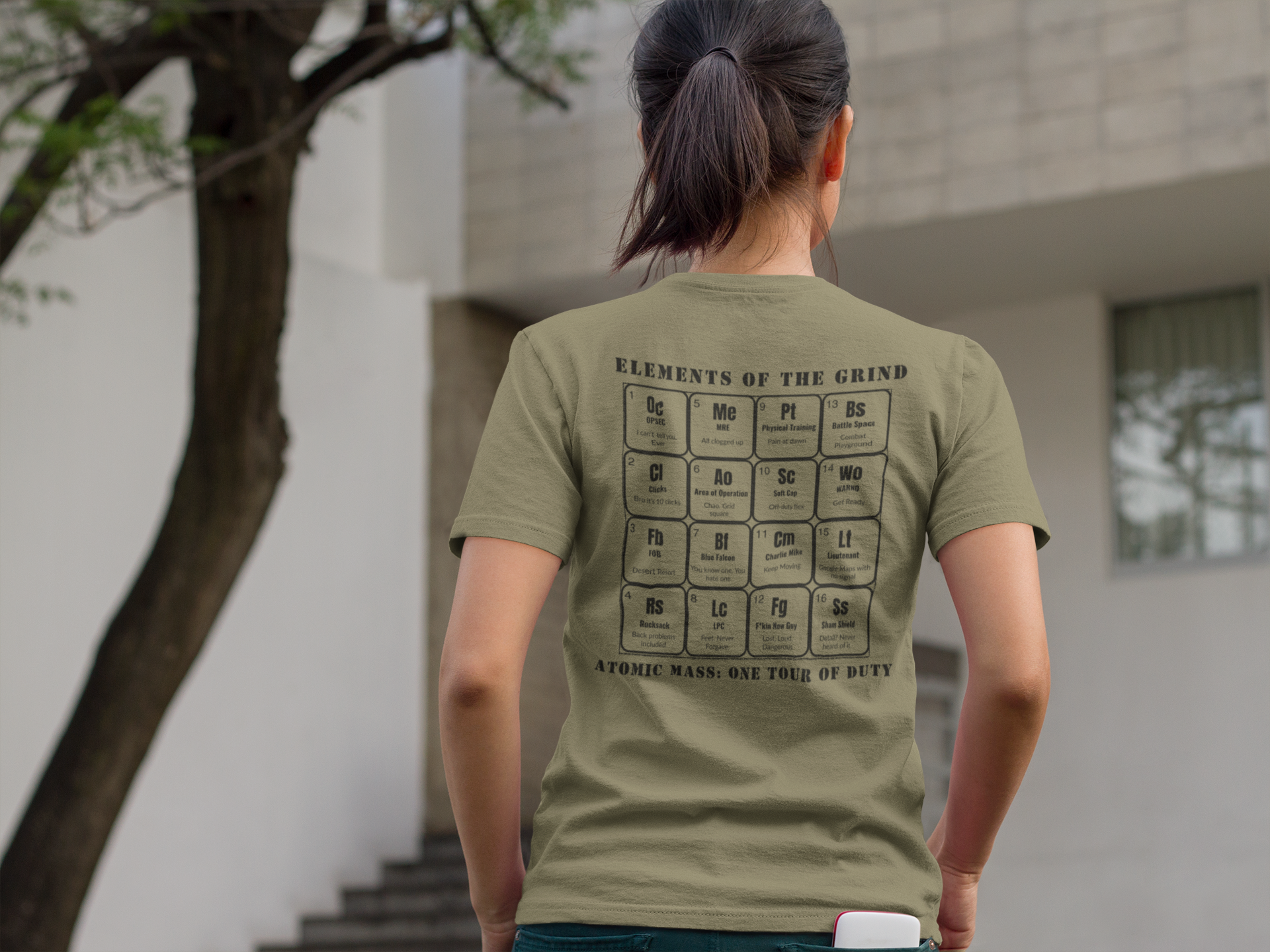 Young woman walking to a building with a phone in back pocket, wearing military green t-shirt, elements of the grind. Tactical pause apparel.
